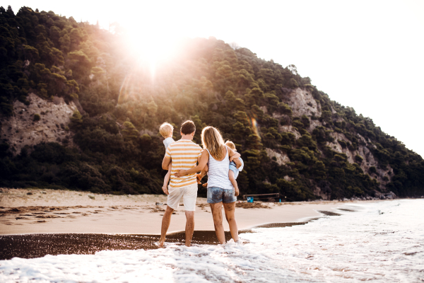 A rear view of family with two toddler children standing on beach on summer holiday at sunset.