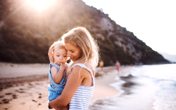 Young mother hugging a toddler girl on beach on summer holiday at sunset.