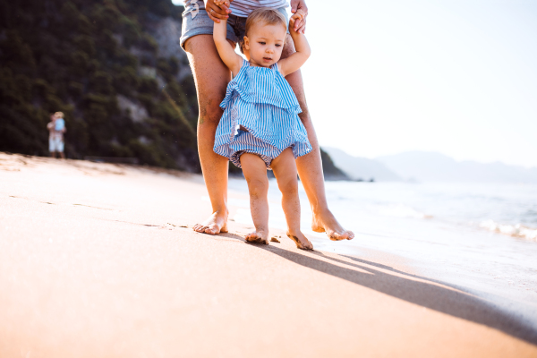 Midsection of young mother walking with a toddler girl on beach on summer holiday at sunset.