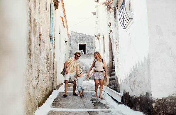 A family with two toddler children walking in town on summer holiday. A father and mother with son and daughter in baby carrier on a narrow street.