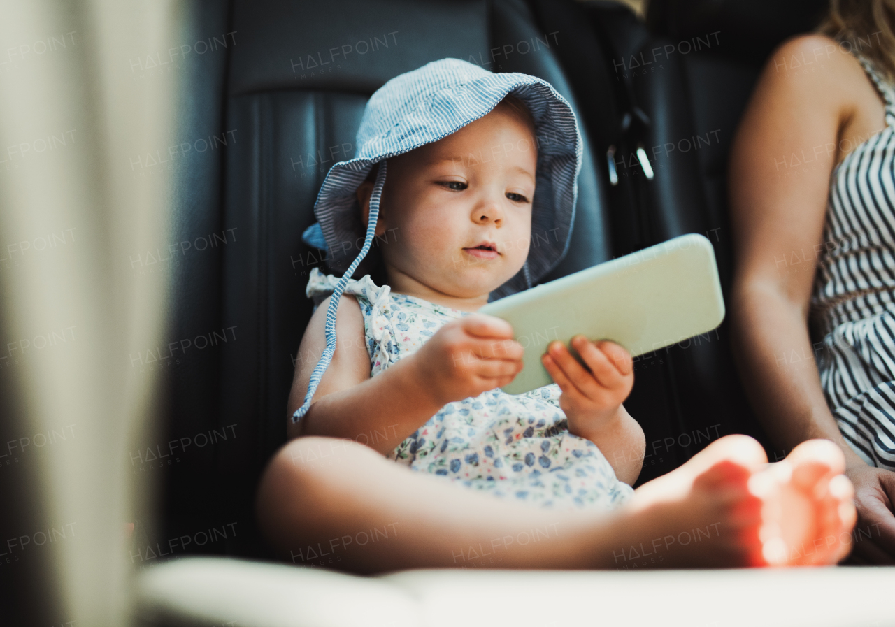 A small toddler girl sitting in a car, playing with smartphone. A summer time.