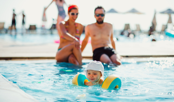 A small toddler child with armbands and parents in swimming pool on summer holiday.