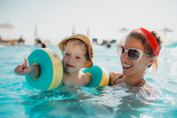 A happy small toddler boy with armbands and his mother swimming in water on summer holiday.