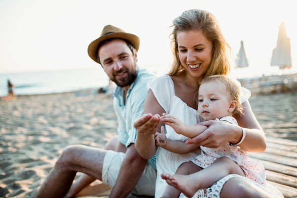 A family with a toddler girl sitting on sand beach on summer holiday, playing.