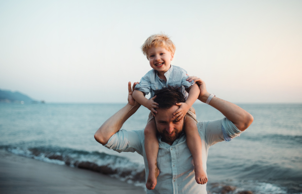 Mature father with a toddler boy standing on beach on summer holiday, having fun.