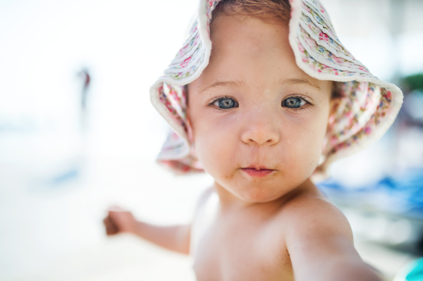 A close-up of cute small toddler girl sitting on beach on summer holiday.