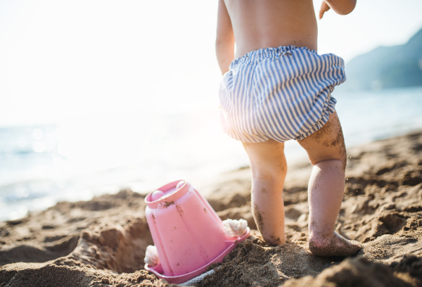 A midsection of small toddler girl with shorts playing on sand beach on summer holiday. A rear view.