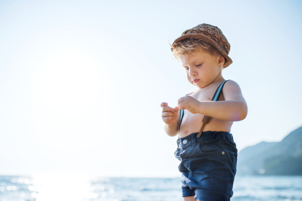 A small toddler boy with hat and shorts standing on beach on summer holiday. Copy space.