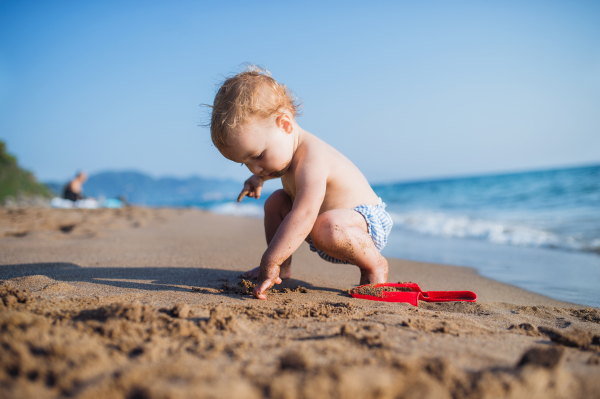 A small topless toddler girl sitting on beach on summer holiday, playing in sand.