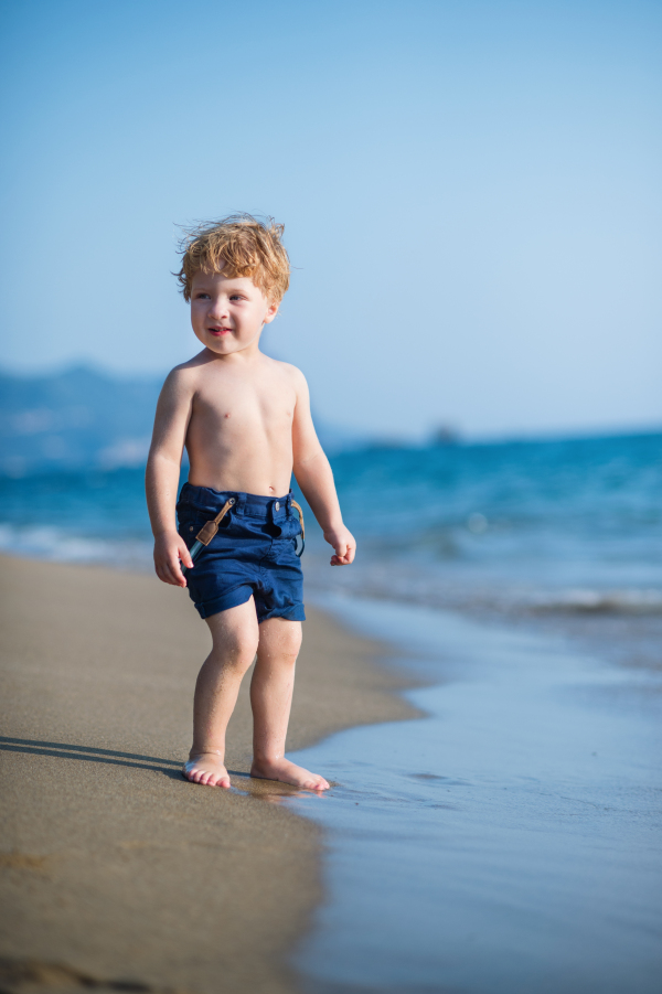 A cute small toddler boy with shorts walking on sand beach on summer holiday. Copy space.