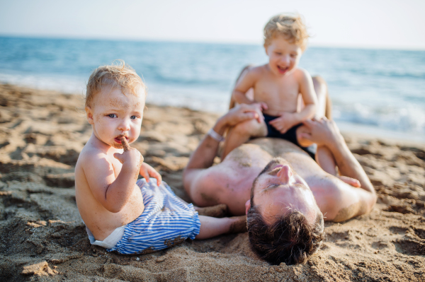 A father with two toddler children lying on sand beach on summer holiday, playing.