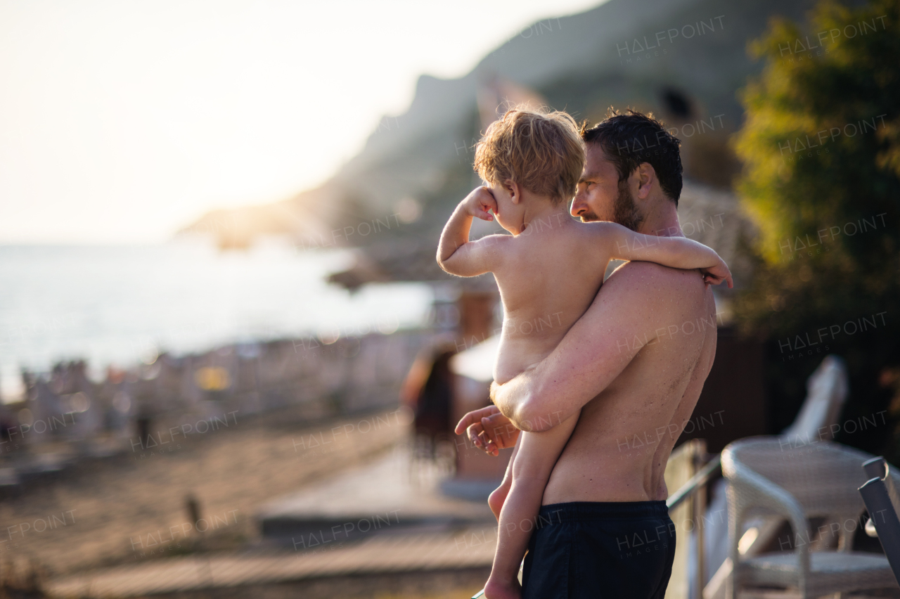 A mature father holding a toddler boy on beach on summer holiday. Copy space.