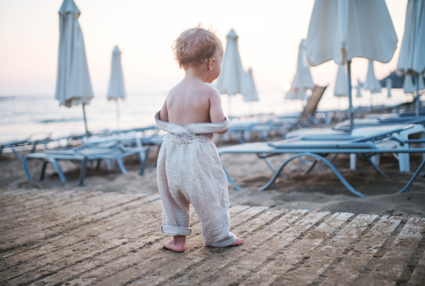 A rear view of small toddler girl walking topless on beach on summer holiday.