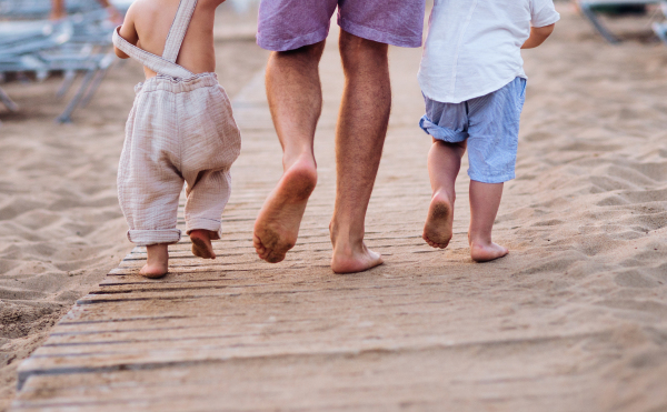 A midsection of father with two toddler children walking barefoot on sand beach on summer holiday. A rear view.