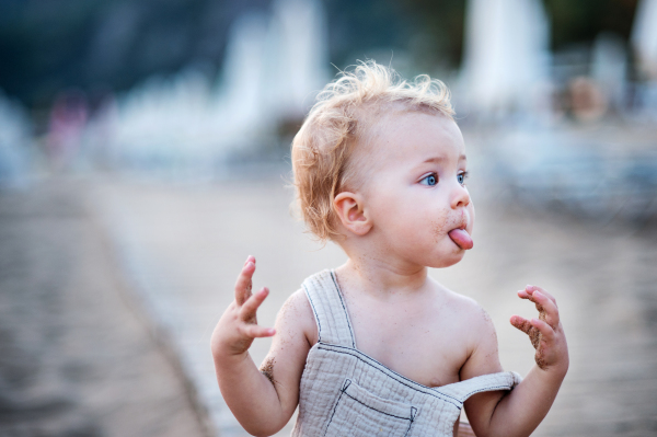 A cute small toddler girl walking on beach on summer holiday, sticking a tongue out.