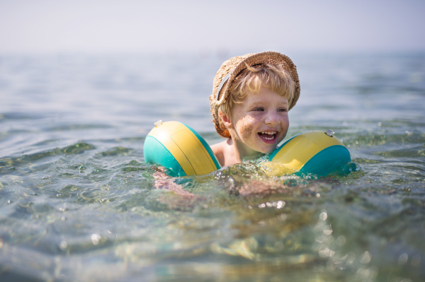 A happy small toddler boy with hat and armbands swimming in water on summer holiday, sand on cheeks.