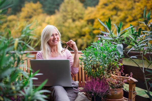 A senior woman with laptop sitting outdoors on terrace, working.