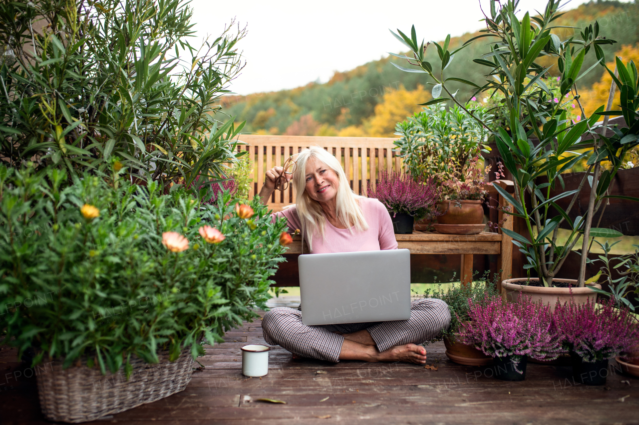 A senior woman with laptop sitting outdoors on terrace, working.