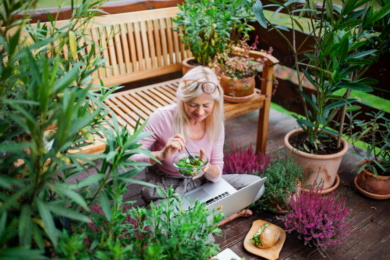 Front view of senior woman with laptop sitting outdoors on terrace, working and eating.