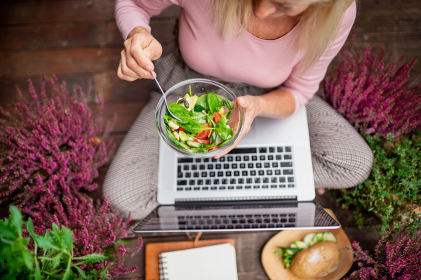 Top view of senior woman with laptop sitting outdoors on terrace,eating healthy lunch.