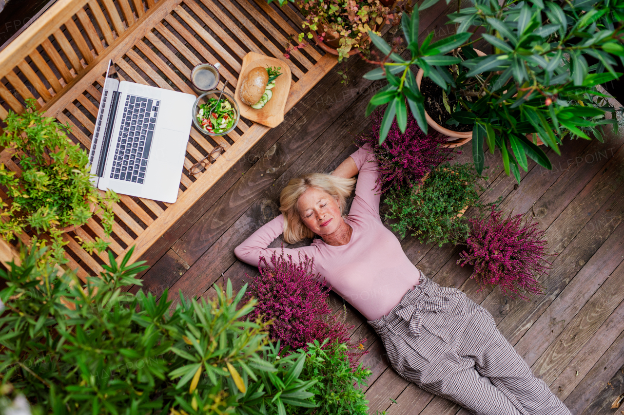 A top view of senior woman with laptop lying outdoors on terrace, resting.