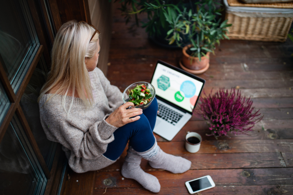 Senior woman with laptop sitting outdoors on terrace, working and eating.