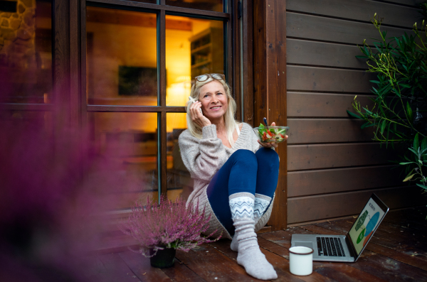 Senior woman with laptop and smartphone sitting outdoors on terrace, working.