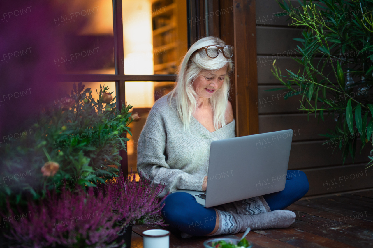 A senior woman with laptop sitting outdoors on terrace, working in the evening.