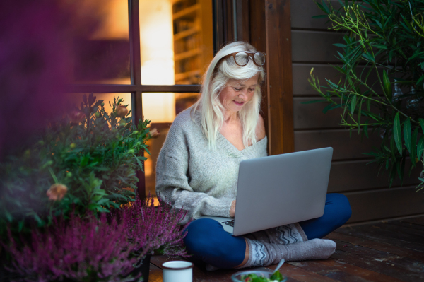 A senior woman with laptop sitting outdoors on terrace, working in the evening.