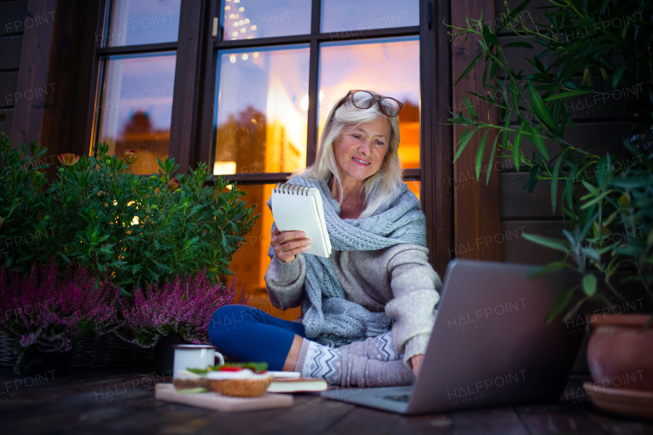 A senior woman with laptop sitting outdoors on terrace, working in the evening.