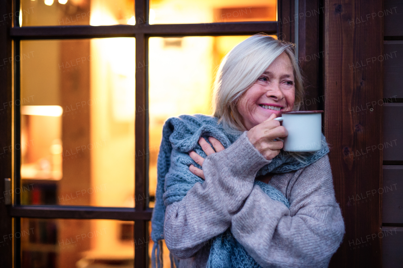 Senior woman with coffee and scarf standing outdoors on terrace, feeling cold.