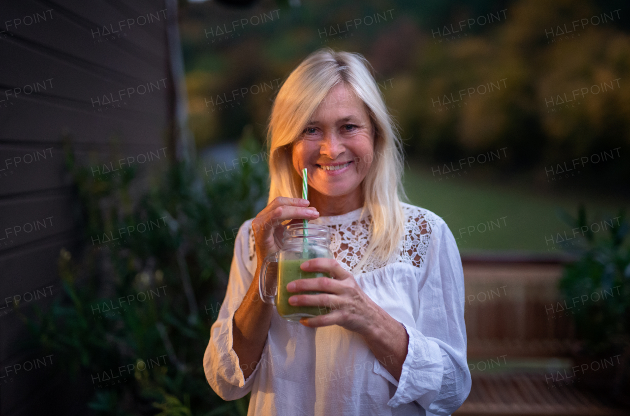 A senior woman with juice standing outdoors on terrace, looking at camera.