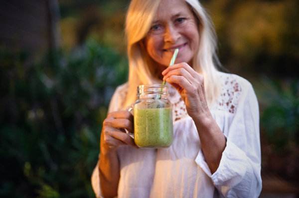 A senior woman with juice standing outdoors on terrace, looking at camera.