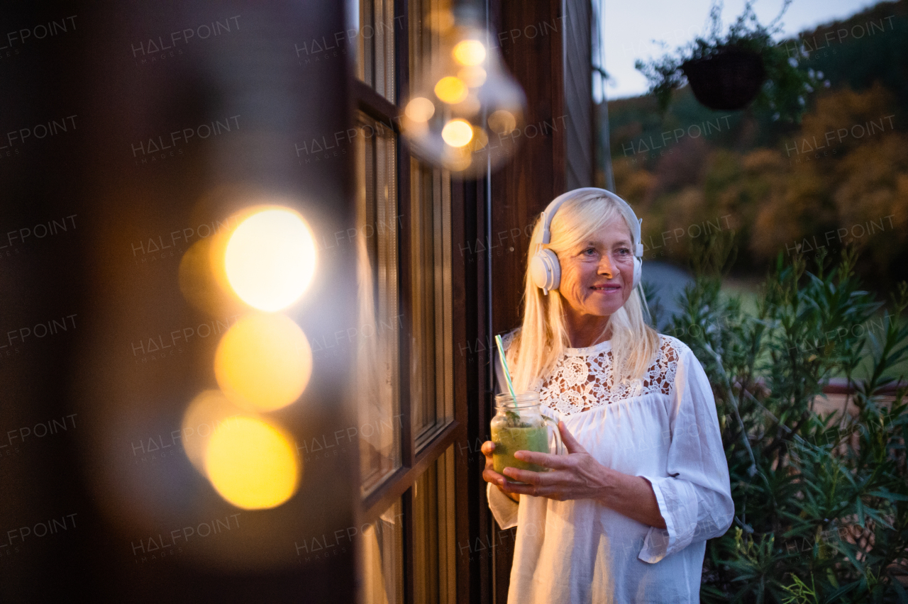 Senior woman with headphones standing outdoors on terrace, drinking smoothie.