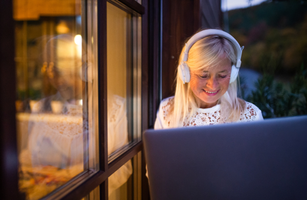 A senior woman with laptop sitting outdoors on terrace, working in the evening.