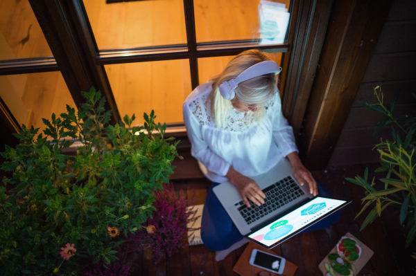 A senior woman with laptop sitting outdoors on terrace, working in the evening.