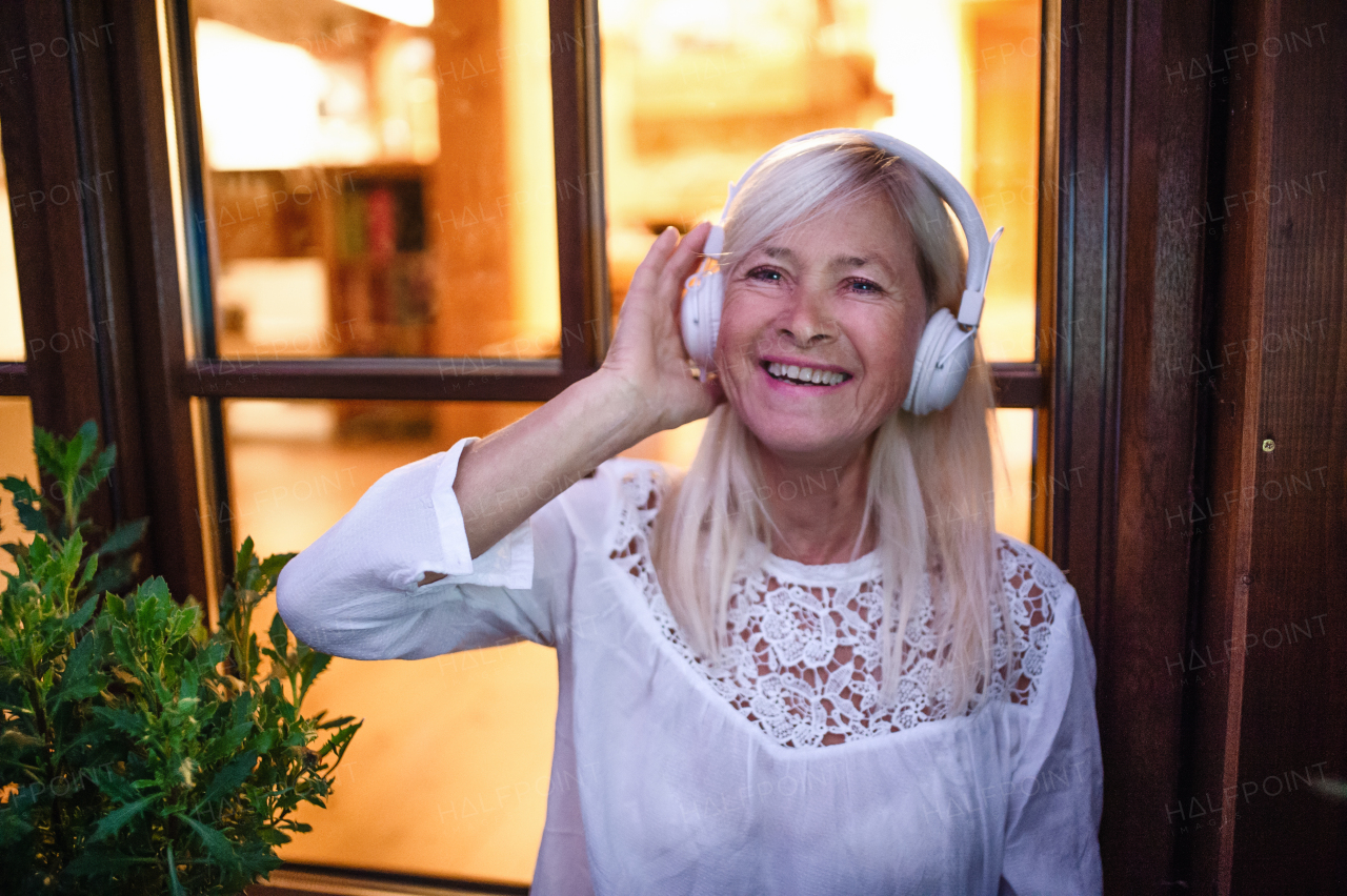 A senior woman with headphones standing outdoors on terrace, listening to music.