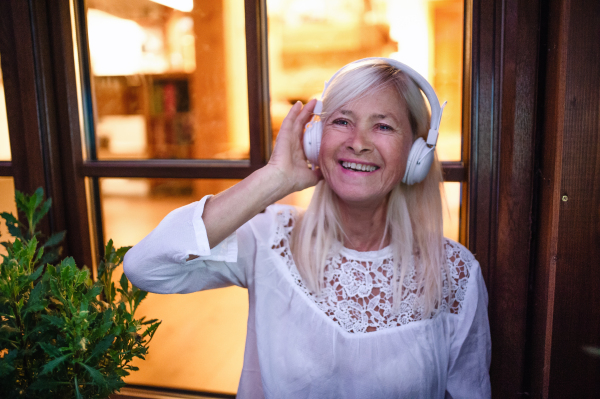 A senior woman with headphones standing outdoors on terrace, listening to music.