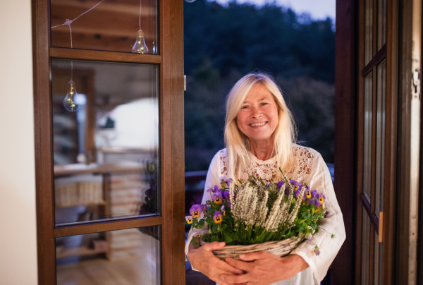 Senior woman with flowers in basket standing outdoors on terrace, entering house.