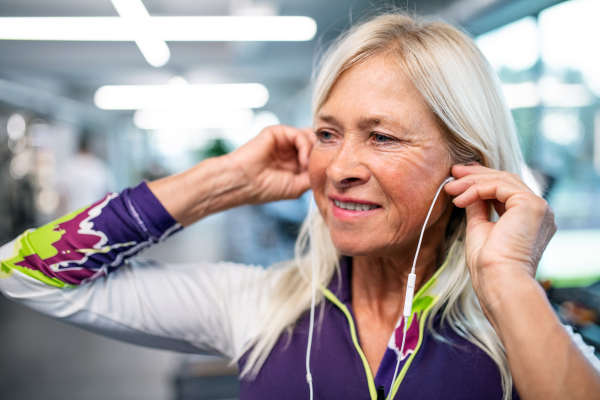 A front view of senior woman with earphones in gym resting after doing exercise.