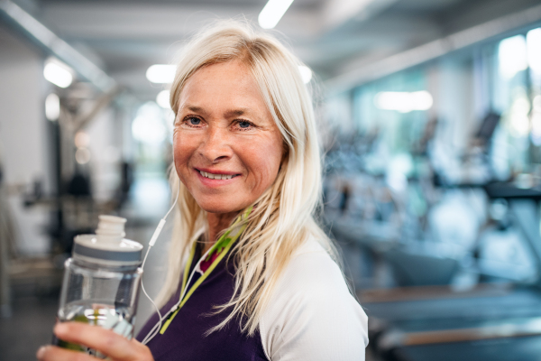 A front view of senior woman with earphones and water bottle in gym, resting after doing exercise.