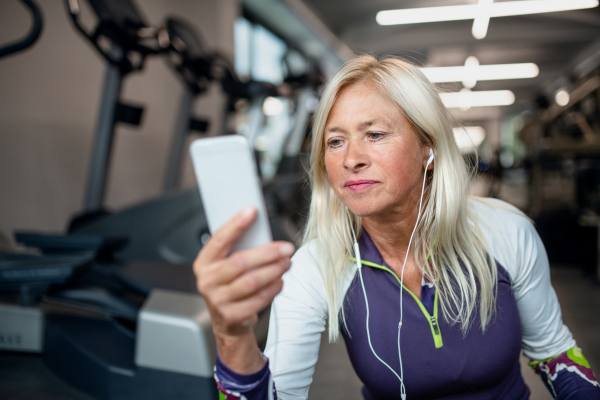 A front view of senior woman with earphones and smartphone in gym resting after doing exercise.