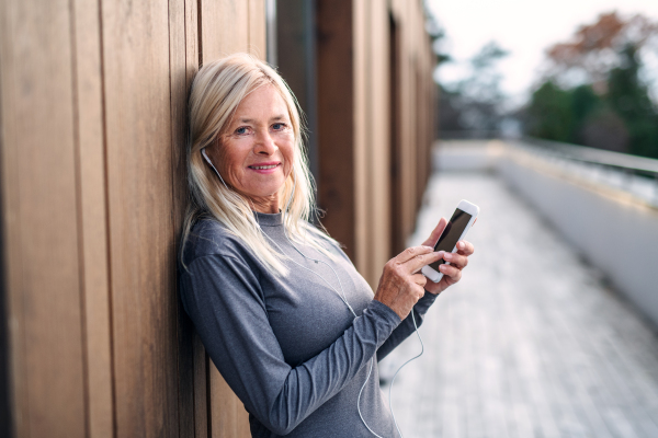 A senior woman with earphones and smartphone outdoors resting after exercise.