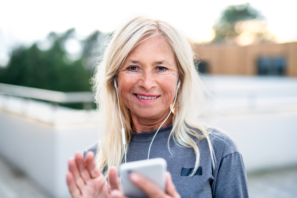 A senior woman with earphones and smartphone outdoors resting after exercise.