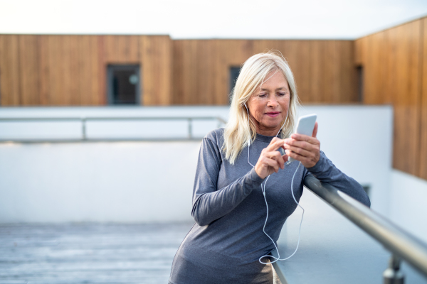 A senior woman with earphones and smartphone outdoors resting after exercise.