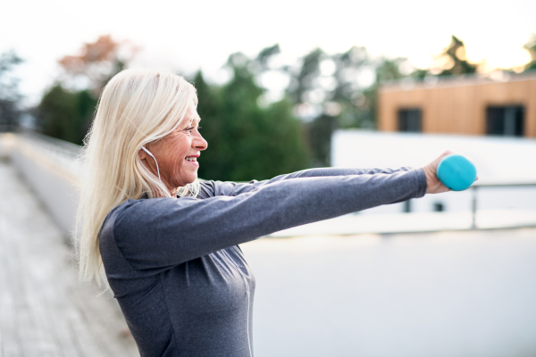 A senior woman with earphones and dumbbells outdoors doing exercise on terrace.