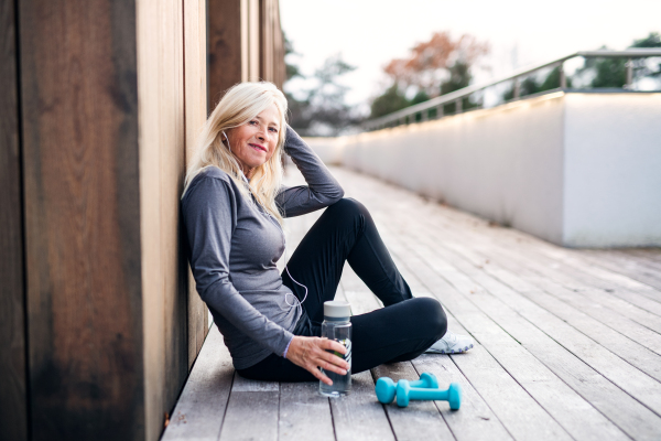 A senior woman with earphones, water bottle and dumbbells outdoors resting after exercise.