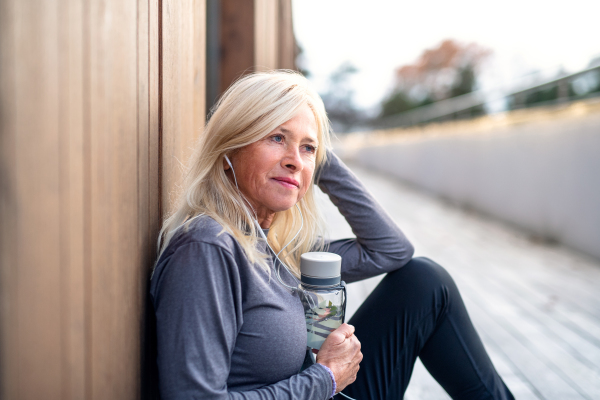 A side view of senior woman with earphones sitting outdoors resting after exercise.