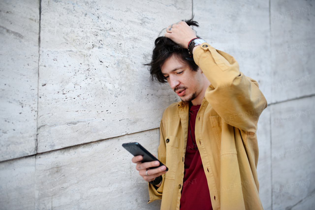 A portrait of young man standing against concrete wall outdoors, using smartphone.