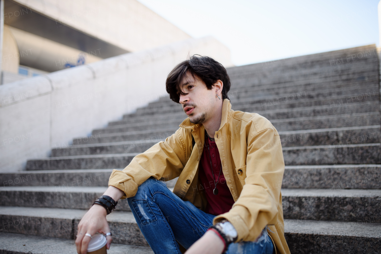 A portrait of sad young man sitting on staircase outdoors in city, resting.